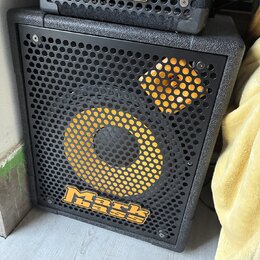 Černý basový reproduktor s kovovou mřížkou, na které je žlutý kruh a čtverec. V dolní části mřížky je umístěno žluté logo Mark bass. Nad ním je vidět…
