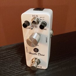 Bílý kytarový efekt s nápisem Hustle Drive. Na horní straně jsou dva černé otočné knoflíky s popisky VOLUME a TONE, přepínač HP/LP a vstup DC IN. Upr…