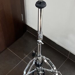 Chromovaná stojka na činely s trojnožkou a pedálem. Na pedálu je viditelný nápis BRUX. Povrch chromu vykazuje známky opotřebení a drobné nečistoty.