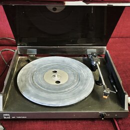 Gramofon s černým šasi a průhledným plastovým krytem, který je otevřený. Na talíři je položena vinylová deska. V levém dolním rohu šasi je bílý štíte…