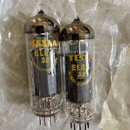 Dvě elektronické vacuumové lampy s průhledným sklem a kovovými patkami. Na obou je žlutý nápis TESLA EL84 32 a u levé lampy je viditelný nápis MADE I…