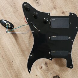 Černý plastový pickguard s třemi integrovanými snímači. Na každém ze snímačů je viditelný nápis emg. Součástí jsou tři černé ovládací knoflíky, přepí…