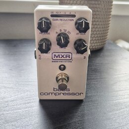 Bílý kytarový pedál s nápisy MXR, BASS INNOVATIONS a bass compressor. Na horní straně je LED indikátor GAIN REDUCTION s hodnotami -20, -10, -7, -5, -…