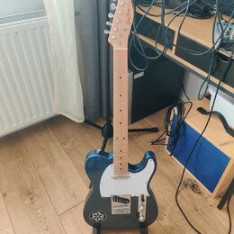 Modrá elektrická kytara s bílým pickguardem a světlou hnedou hmatníkovou deskou. Na hlavě je vidět nápis „Moody Blues“. Kytara stojí na černém stojan…