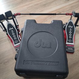 Červený dvojpedál na bubny s kovovými šlapkami. Na obou šlapkách je bílý nápis DW 5000 a The Drummer's Choice. Pedál je umístěn za černým plastovým k…