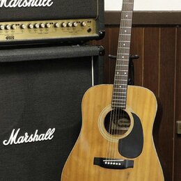 Akustická kytara s přírodním dřevem a černým plektrem. Na hlavě hmatníku je viditelné logo Yamaha. Vedle kytary stojí černý zesilovač a reproduktorov…