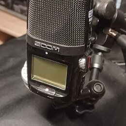 Černý digitální záznamový přístroj značky ZOOM s kovovou mřížkou mikrofonů v horní části. Zařízení má malý LCD displej a je připevněno na černý stati…