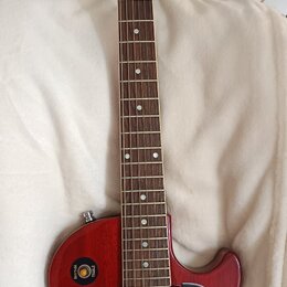 Červená elektrická kytara s tmavým hnedým hmatníkem. Na hlavě je bílé logo Gibson a nápis Les Paul. Černý plechtrguard obsahuje několik podpisů zlato…