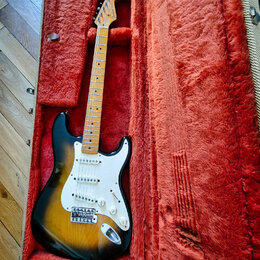 Elektrická kytara s tónem sunburst a bílým pickguardem v otevřeném kufru s červenou plišovou výstelkou. Na hlavě hředu je viditelné logo Fender. Kyta…