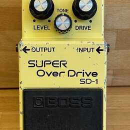 Žlutý kytarový efekt s nápisy SUPER Over Drive a SD-1. Na horní straně jsou tři ovládací knoflíky s popisky LEVEL, TONE a DRIVE a kontrolka CHECK. V…
