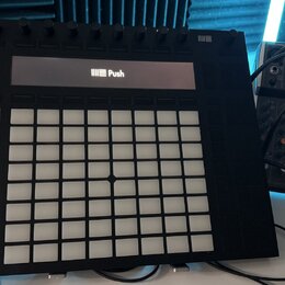 Černý kontrolér Ableton Push s mřížkou bílých podsvícených polí a řadou otočných ovladačů v horní části. Na displeji je vidět logo a nápis Push. Vedl…