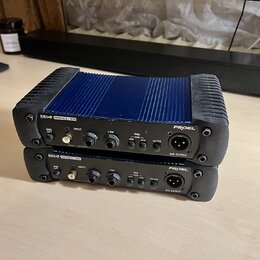 Dvě pasivní DI boxy značky PROEL, model DB1+P PASSIVE D.I.BOX. Horní kus má modré kovové tělo s žebrovaným povrchem a černé plastové kryty na koncích…