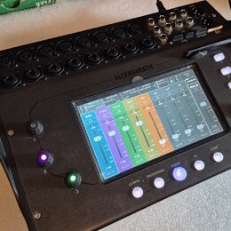 Černý digitální mixovací pult s dotykovým displejem, na kterém je zobrazeno míchací rozhraní s barevnými faderem. Na horní straně je nápis ALLEN&HEAT…