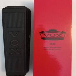 Černý wah-wah pedál s texturou na povrchu a vyraženým logem VOX. Vedle něj je červená papírová krabice s logem VOX AMPLIFICATION LTD. a nápisy V845,…