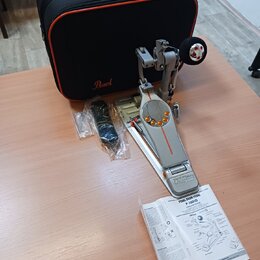 Na fotografii je stříbrný bubnový pedál s oranžovými detaily a nápisem IDemon. Vedle něj leží černý přepravní kufr s logem Pearl, náhradné části v pl…