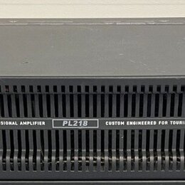 Černý rackový zesilovač s nápisy QSC POWERLIGHT 2, 1800 WATT PROFESSIONAL AMPLIFIER a PL218 CUSTOM ENGINEERED FOR TOURING. Na pravé straně jsou dva o…