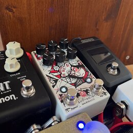Detailní záběr na několik kytarových efektů v pedalboardu. Uprostřed je bílý pedál s červenobílou grafikou, který nese nápisy ratio, program a v doln…