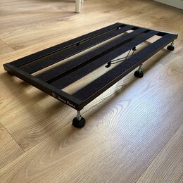 Černý kovový pedalboard s mřížkovým povrchem z gumových nebo plstěných pásů. Na boční hraně je bílý nápis „Harley Benton“ s logem. Nástroj stojí na č…