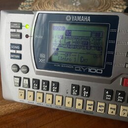 Stříbrný sekvenční modul s displejem a klávesovou ovládací. Na horní straně je logo YAMAHA a pod displejem nápis MUSIC SEQUENCER QY100. V levé části…