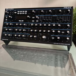 Černý syntetizátor s nápisy novation a PEAK. Na spodním okraku je text OXFORD-OSCILLATORS a EIGHT-VOICE POLYPHONIC SYNTHESISER. Přístroj obsahuje num…