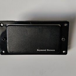 Černý kytarový snímač s krytem. V pravém dolním rohu je bílý nápis Seymour Duncan. Snímač je umístěn v černé rámečku s otvory pro šrouby. Povrch kryt…
