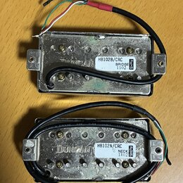 Dva kytarové humbuckery s kovovým krytem a připojenými kabely. Horní snímač má štítek s nápisy HB102B/CRC, BRIDGE a 1102. Dolní snímač má štítek s ná…