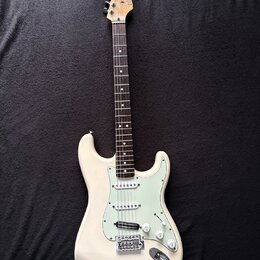 Bílá elektrická kytara typu Stratocaster s jasně zeleným pickguardem. Na hlavě hředu je viditelné logo Fender. Nástroj vykazuje známky používání, jso…