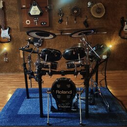 Elektronická bicí souprava značky Roland s černými bubny a činely. Na basovém bubnu je bílý nápis Roland a V-Drums. Souprava je umístěna na černém ko…