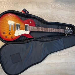 Elektrická kytara s červeně-žlutým sunburst povrchem těla a bílým pickguardem, položená v otevřeném černém softcase. Na hlavě kytary je viditelné log…