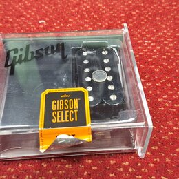 V průhledném plastovém boxu se nachází černý elektrický kytarový snímač. Na povrchu je vidět bílé logo Gibson a žlutá nálepka s nápisem GIBSON SELECT…