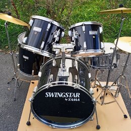 Černá bubnová sada s kovovým kováním a zlatými činely. Na basovém bubnu, tom-tamech i snare je viditelný nápis SWINGSTAR by TAMA. Na jednom z činelů…