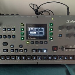 Stříbrný sampler s nápisem Elektron a Octatrack MKII. Zařízení obsahuje displej, několik ovládacích knoflíků, tlačítka s různými funkcemi a v dolní č…