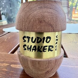 Dřevěný shaker s kovovým středovým prutem zlaté barvy. Na kovové části je černý nápis STUDIO SHAKER a svisle orientovaný text SMALL. Povrch dřeva vyk…