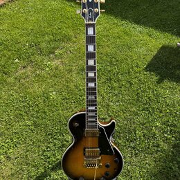 Elektrická kytara s tónem těla v barvě sunburst a černým pickguardem. Na hlavě je viditelné logo Gibson a nápis Custom. Hриф má vložky z perlemi a na…