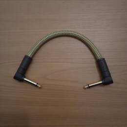 Kabel s úhlovými 6,3mm konektory v barvě zlata. Kabel má pletený obal v béžové barvě a černé gumové chrániče u konců. Na jednom z chráničů je nečitel…