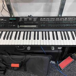 Černý digitální klavír s bílými a černými klávesami. Na levé straně je nápis YAMAHA DX7FD. Nástroj je umístěn v černém transportním kufru s kovovými…