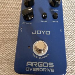 Modrý kytarový efekt s nápisy JOYO a ARGOS OVERDRIVE. Na horní straně jsou tři velké černé otočné knoflíky s označením ATTACK a LEVEL, dvě menší čern…