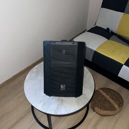 Černý reproduktor s mřížkou v horní části a logem "E" ve spodní části. Nástroj stojí na kulatém stole s mramorovým povrchem a černou kovovou nohou. P…