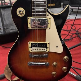 Elektrická kytara s tmelem v barvě sunburst (černá přecházející do hnědé). Má dva humbuckery s kovovými kryty, bílý pickguard a čtyři zlaté ovládací…