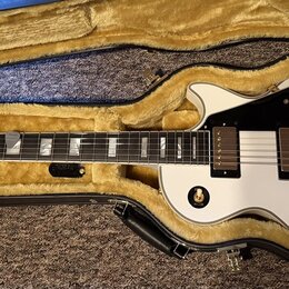 Bílá elektrická kytara s tvarem single-cut v černém kufru s žlutým vnitřkem. Na hlavě je logo Epiphone a nápis "Elite". Kytara má dva humbuckery, zla…