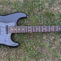 Černá elektrická kytara s šedým mramorovaným pickguardem a hnědým hmatníkem. Na hlavě je viditelné logo Fender a nápis STRATOCASTER, níže je drobný n…