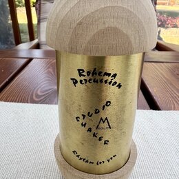 Kovový shaker zlaté barvy s horní a spodní částí z přírodního světlého dřeva. Na kovovém těle jsou černým nápisy „Rohema Percussion“, „Saloio SHAKER“…