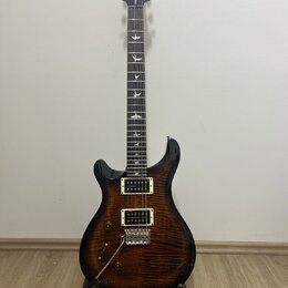 Elektrická kytara s tmavě hnědým plamenovitým povrchem těla a černou hlavou. Na hlavě je bílé logo PRS a nápis SE Custom. Hmatník obsahuje bílé vložk…