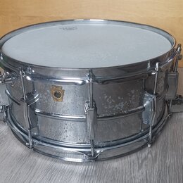 Kovový snare drum s chromovaným povrchem a bílým bubenovou skins. Na korpusu je viditelné zlaté logo ve tvaru štítu, ale text uvnitř něj není čitelný…