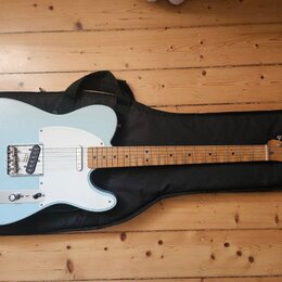 Světle modrá elektrická kytara typu Telecaster s bílým pickguardem, položená na černém softcasu. Na hlavě hředu je viditelné logo Fender a nápis MADE…