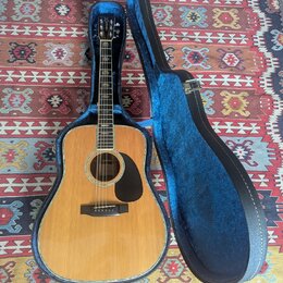 Akustická kytara s přírodním dřevem a černým plechtrem v modrém tvrdém pouzdře. Na hlavě nástroje je viditelné logo "Epiphone". Kytara i pouzdro vyka…