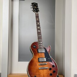 Elektrická kytara s tvarem singlecut a povrchovou úpravou v hnědém sunburstu. Na hlavě je viditelné logo Gibson. Kytara má dva humbuckery, čtyři otoč…