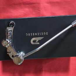 Chromovaný kytarový tremolo systém položený na černé krabici. Na krabici je bílý nápis DUESENBERG a logo ve tvaru stylizovaného písmene D s křídly. P…
