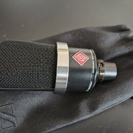 Černý kondenzátorový mikrofon s kovovou síťovkou a stříbrným prstencem, položený na černém látkovém vaku. Na těle mikrofonu je červenobílý kosočtverc…
