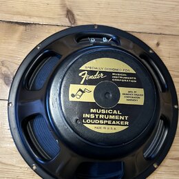 Černý reproduktor s kovovým rámem, položený na dřevěném povrchu. Na zadní straně je žlutá nálepka s nápisy „SPECIALLY DESIGNED FOR Fender MUSICAL INS…